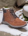 03610 Lace-Up Ankle Boot in brown leather with contrast dark trim, photographed on snowy surface. Features moc-toe design, rugged lacing system, and gray rubber sole. Water-repellent winter boot shown in outdoor setting with snow and moss accents.