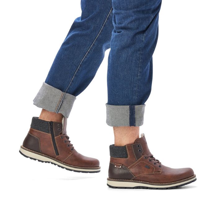 Legs in cuffed jeans wearing brown ankle boot with textile cuff, laces, white midsole and brown outsole.