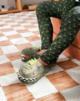 Person wearing dinosaur slippers and green scale-patterned pants sits on a bed on a checkered carpet.