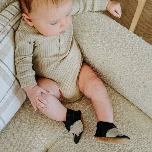 Baby wearing Robeez Soft Sole Shoes - Friends in black with suede soles, sitting on a beige couch. Ideal for toddlers, promoting balance and growth.