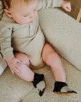 Baby wearing Robeez Soft Sole Shoes - Friends in black with suede soles, sitting on a beige couch. Ideal for toddlers, promoting balance and growth.