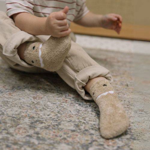 Toddler wearing beige dinosaur-themed cozy socks on a textured rug.