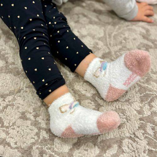 Toddler wearing white unicorn-themed cozy socks on a patterned rug.