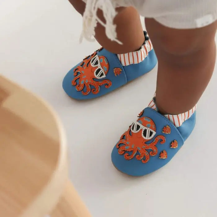 Baby wearing blue leather shoes with orange octopus and fish on them.