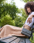 Brunette wearing white and beige skirt outfit sitting on park bench wearing the Addison gray bag as a crossbody