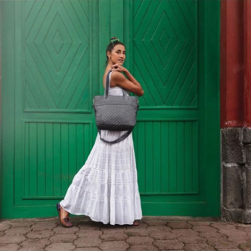 Girl in White Dress standing side view on cobble stone in front of big green doors holding a Travelon Gray Heather Tote bag on shoulder.