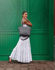 Girl in White Dress standing side view on cobble stone in front of big green doors holding a Travelon Gray Heather Tote bag on shoulder.