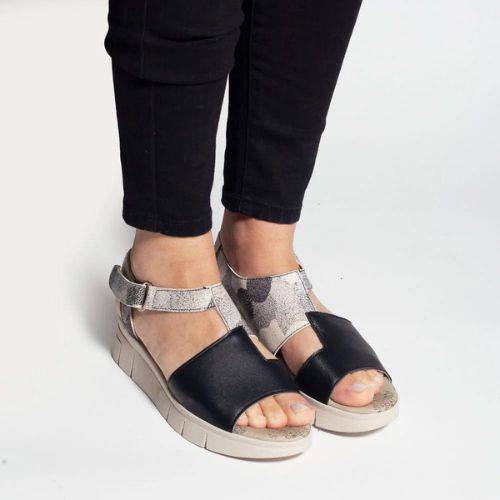 Women in black jeans wearing the Wedge sandal with grey and black leather upper and off white wedge outsole. Asymmetrical upper features adjustable ankle strap.