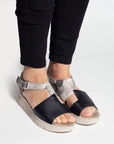 Women in black jeans wearing the Wedge sandal with grey and black leather upper and off white wedge outsole. Asymmetrical upper features adjustable ankle strap.