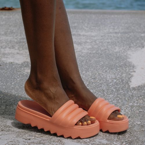 Woman on the street wearing the Pool Party Slide Sandals in Coral. She has matching colour painted toenails. 