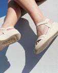Woman sitting on a white wall wearing the Flowt LX Wedge sandal. Picture taken from below. 