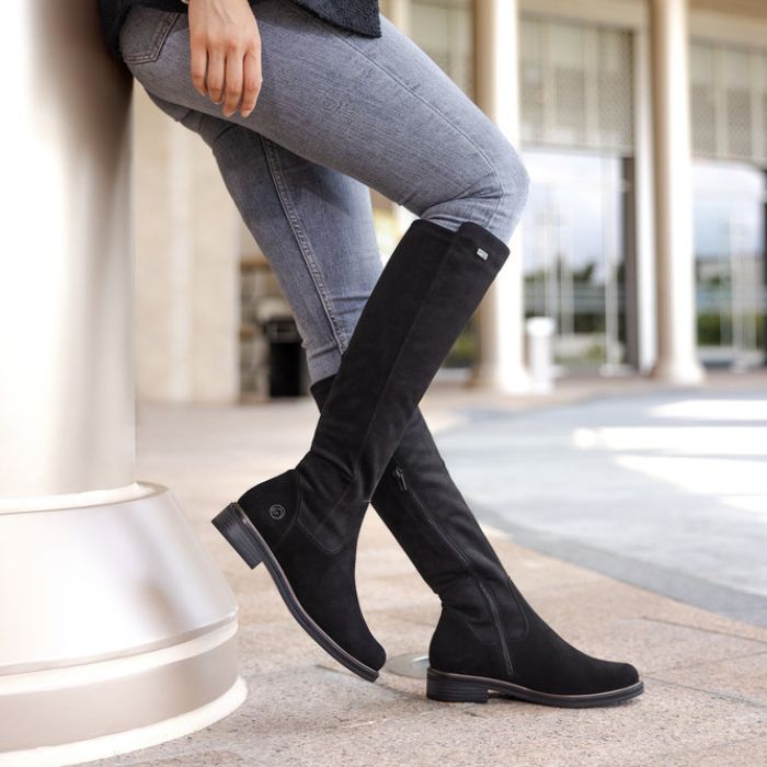 Legs in jeans wearing black tall boot with inside zipper and small silver Remonte emblem at top.