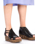 Close-up of a person wearing platform wedge sandals, featuring a cork sole, cut-out design, and side zipper. 