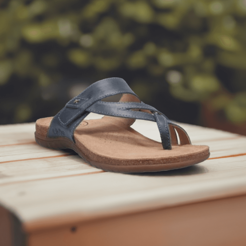 Blue Taos Perfect Slide Sandal displayed outdoors on a wooden surface, featuring leather straps and cushioned footbed. 