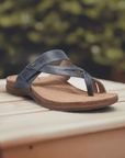 Blue Taos Perfect Slide Sandal displayed outdoors on a wooden surface, featuring leather straps and cushioned footbed. 