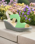 Yoxa Wedge Sandal in green displayed outdoors with floral background, highlighting its stylish design and comfort.