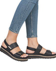 Legs of woman in blue jeans wearing black backstrap sandals with gold decorated bars on straps.