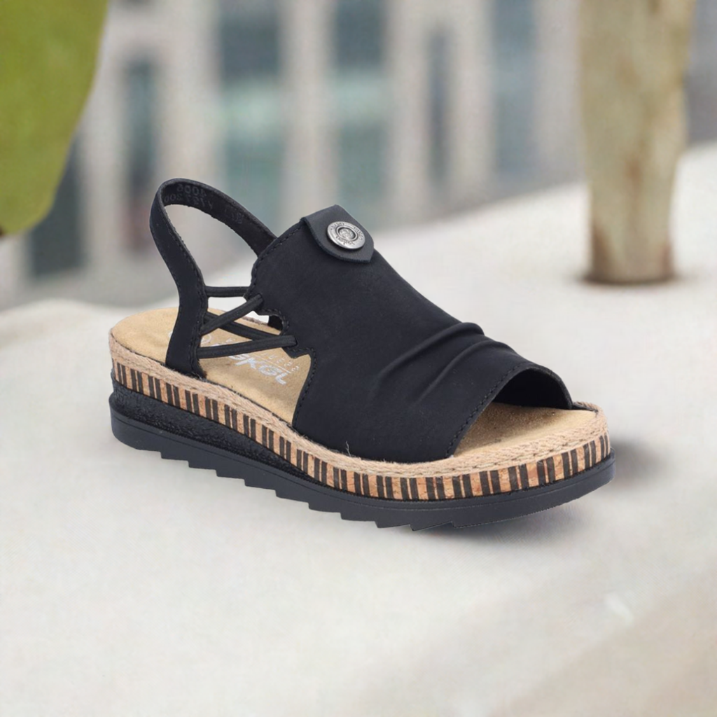 A scenic displayed black Rieker sandal with small gathering on upper, heel strap with criss-cross design at sides, beige twine midsole and black rubber outsole.