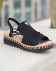 A scenic displayed black Rieker sandal with small gathering on upper, heel strap with criss-cross design at sides, beige twine midsole and black rubber outsole.