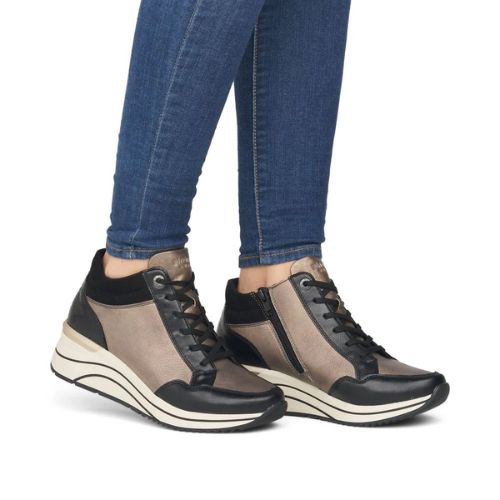 Woman in blue jeans wearing gold and black lace-up wedge sneakers. The left heel is slightly lifted