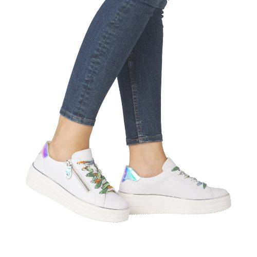 Legs of woman in jeans wearing all white platform sneakers with iridescent heel cap and green/yellow laces.