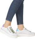 Legs of woman in jeans wearing all white platform sneakers with iridescent heel cap and green/yellow laces.