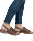 Woman in blue jeans wearing brown leather backstrap sandal with perforated upper and brown outsole.