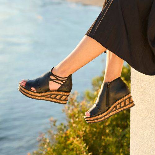 A person in a black skirt sits on a ledge wearing black platform wedge sandals with cut-out designs and cork soles.