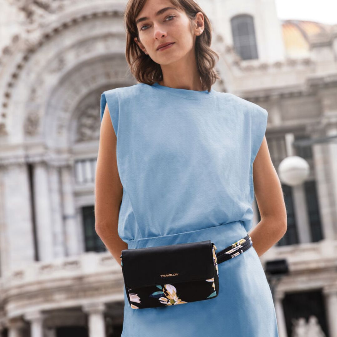 Women in blue dress wearing a black belt bag with floral print. Gold Travelon logo emblem is on the front flap.