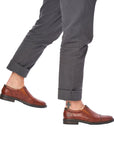 Man in grey pants wearing brown slip on dress shoes with Rieker logo on heel.